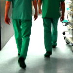 Rückansicht von zwei Personen in grüner OP-Kleidung (Kasack und Hose), die einen hell erleuchteten Krankenhausflur entlanggehen