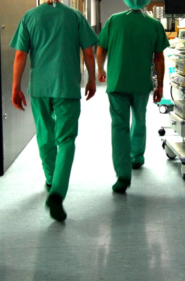 Rückansicht von zwei Personen in grüner OP-Kleidung (Kasack und Hose), die einen hell erleuchteten Krankenhausflur entlanggehen