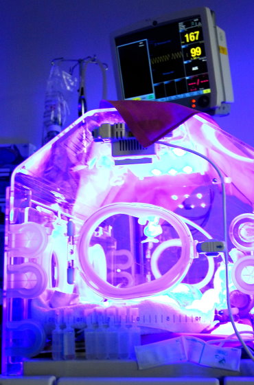 Ein medizinischer Inkubator für Frühgeborene im Einsatz. Der Innenraum wird durch eine Fototherapie-Lampe intensiv violett-blau beleuchtet. Im Hintergrund ist ein Monitor zur Überwachung der Vitalwerte erkennbar.