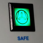 Nahaufnahme einer grün leuchtenden Kontrollanzeige mit einem dreiblättrigen Symbol (Ventilator/Lüfter). Über der Anzeige steht das Wort „SICHER“ und darunter „SAFE“.