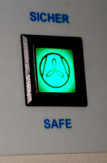Nahaufnahme einer grün leuchtenden Kontrollanzeige mit einem dreiblättrigen Symbol (Ventilator/Lüfter). Über der Anzeige steht das Wort „SICHER“ und darunter „SAFE“.